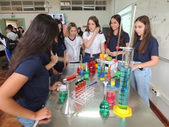 A foto mostra um grupo de estudantes realizando uma atividade experimental com líquidos coloridos em diferentes recipientes de vidro, como provetas e tubos de ensaio. Os estudantes parecem estar concentrados na experiência, e há uma variedade de equipamentos sobre a mesa, incluindo vidrarias e frascos com líquidos de diversas cores. Ao fundo, é possível ver uma lousa branca e outras pessoas no ambiente.